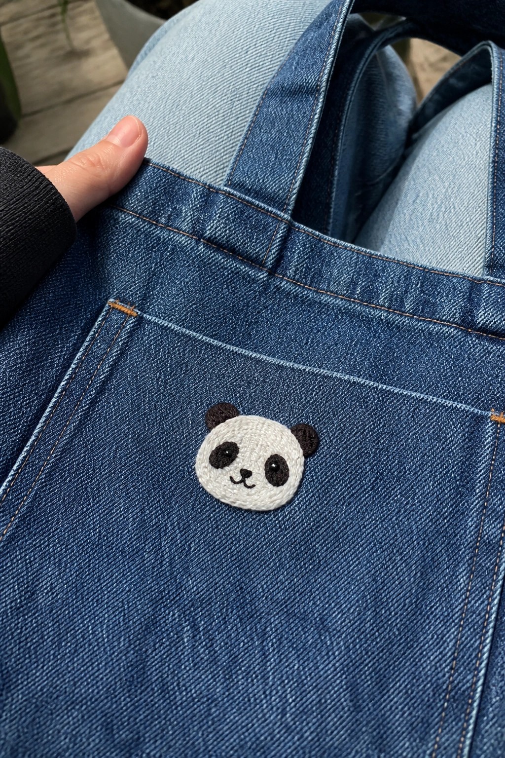 Blue denim overalls pocket featuring embroidered panda face with hand gripping edge