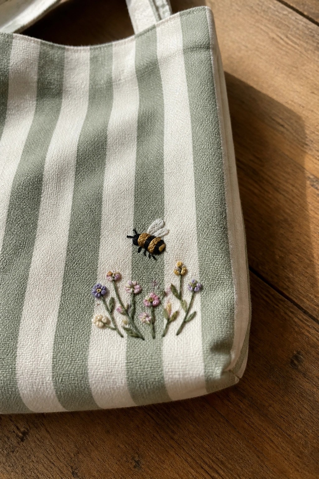A green and white striped tote bag with an embroidered bee above a row of small colorful flowers.