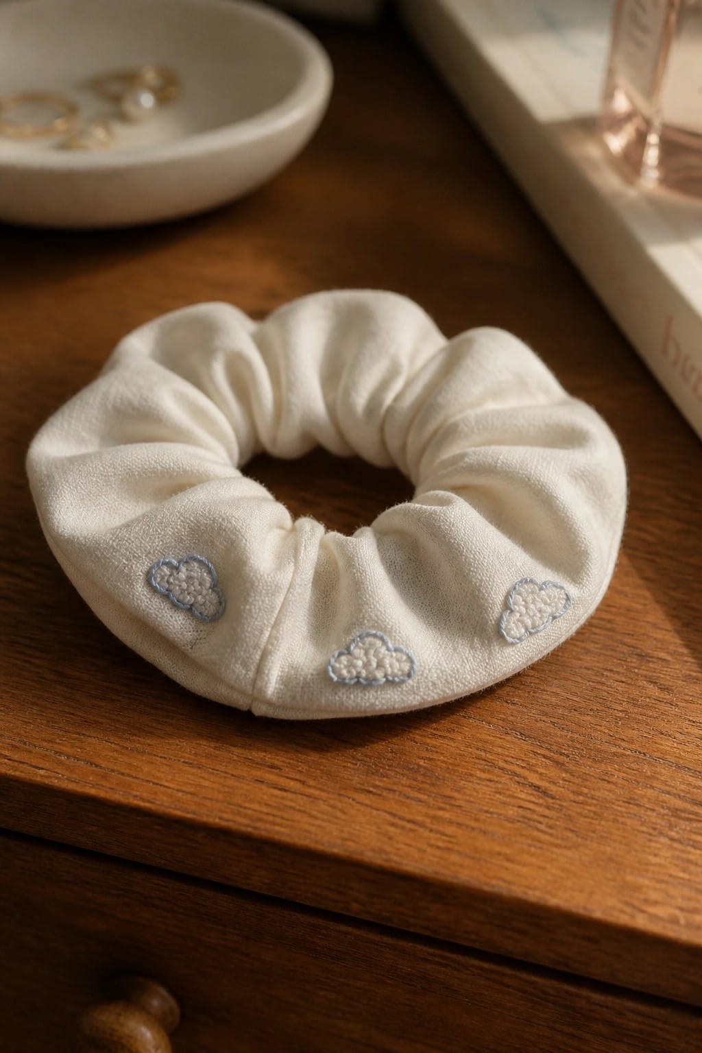 A white scrunchie with three small embroidered clouds spaced around it rests on a wooden surface.