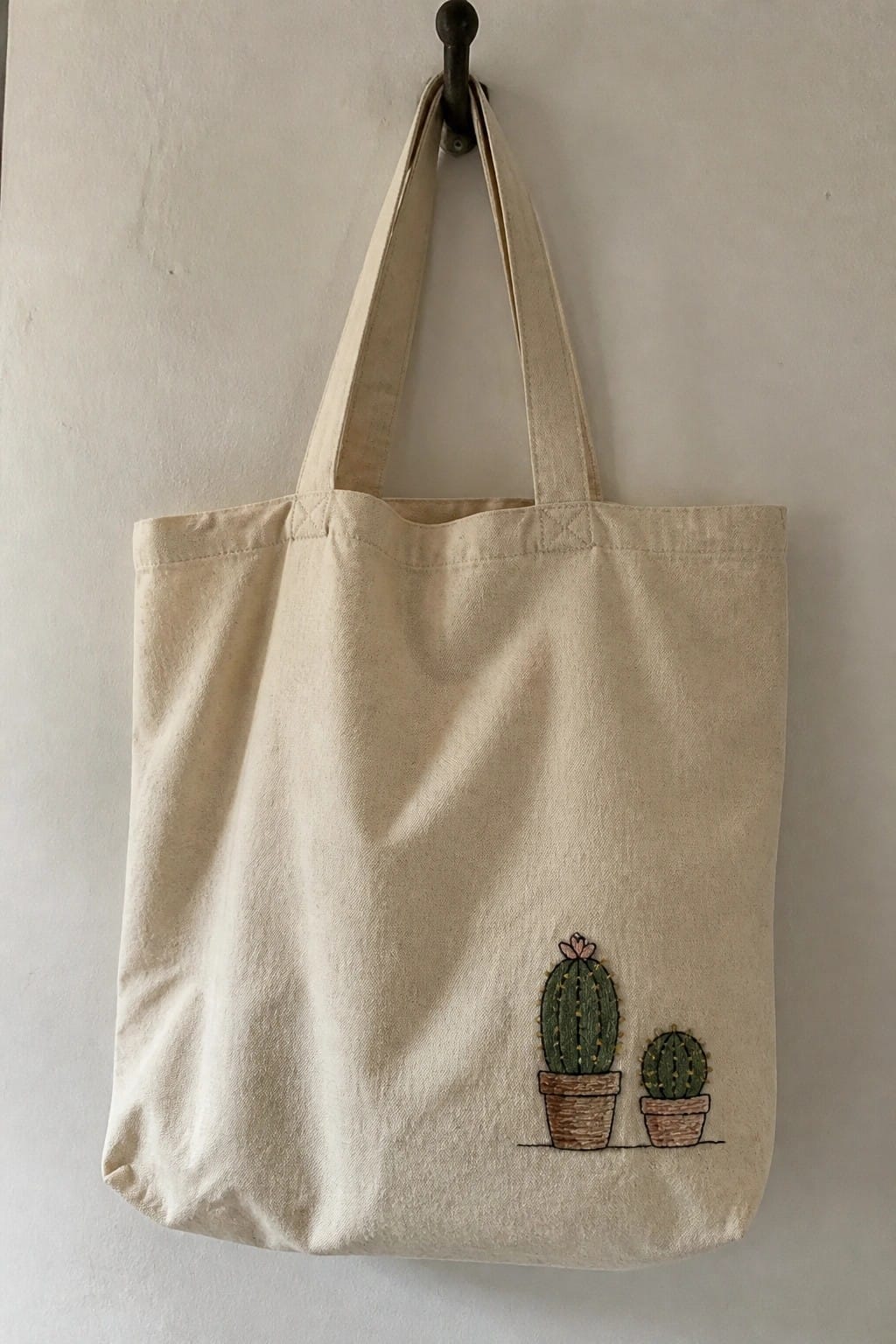 A small off-white zippered pouch with a green embroidered cactus in an orange pot rests on floral fabric.