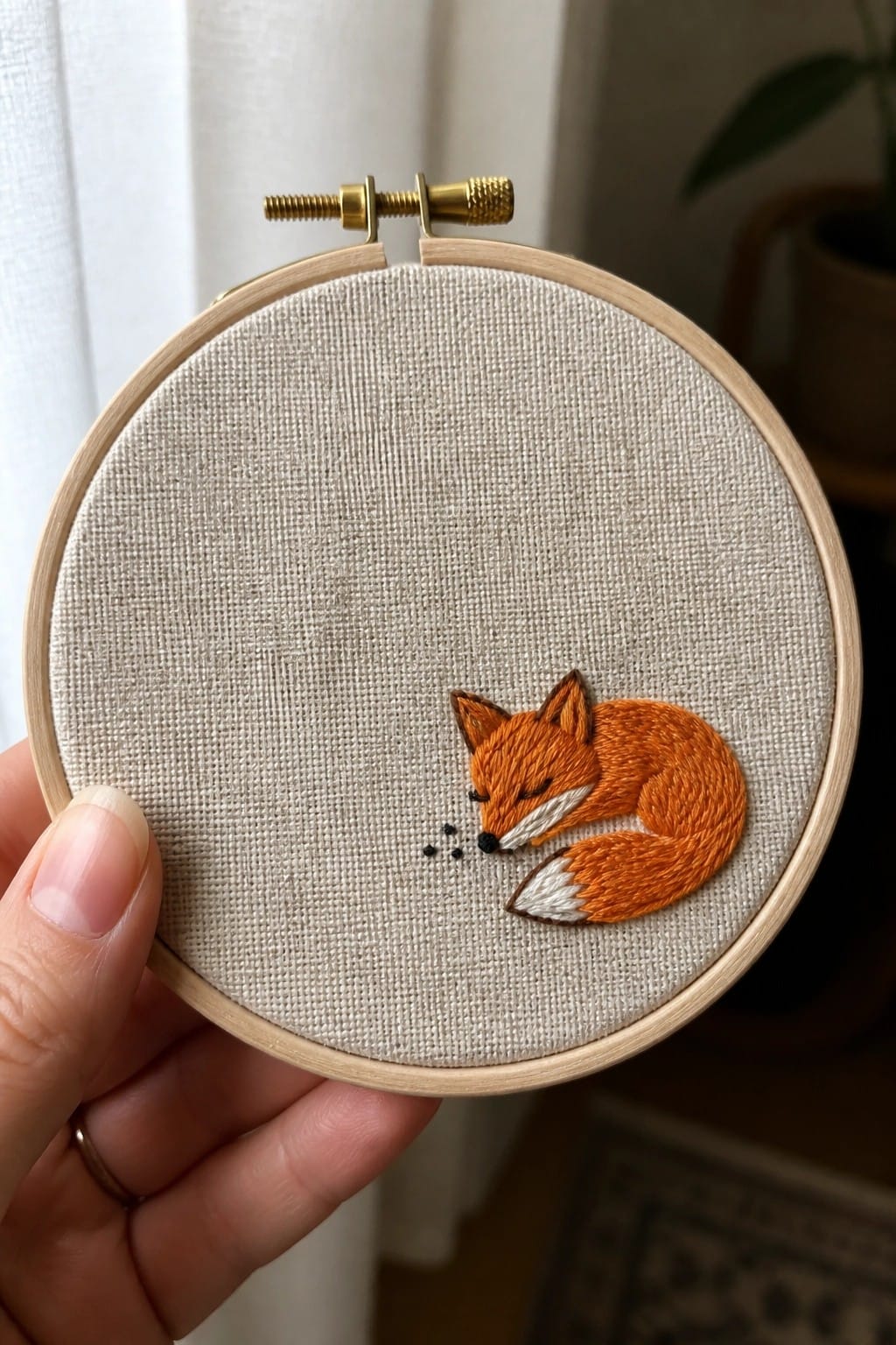 Hand in cream sweater holding hoop with embroidered sleeping orange fox on beige fabric.