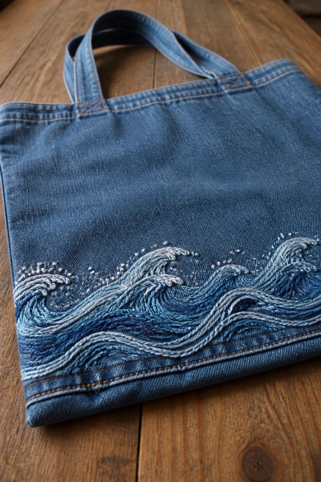 A blue denim tote bag with embroidered waves in shades of blue and white thread along the bottom resting on a wooden table.