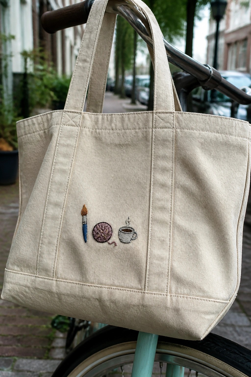 Beige canvas tote bag embroidered with paintbrush, yarn, and coffee cup on bicycle handlebars.