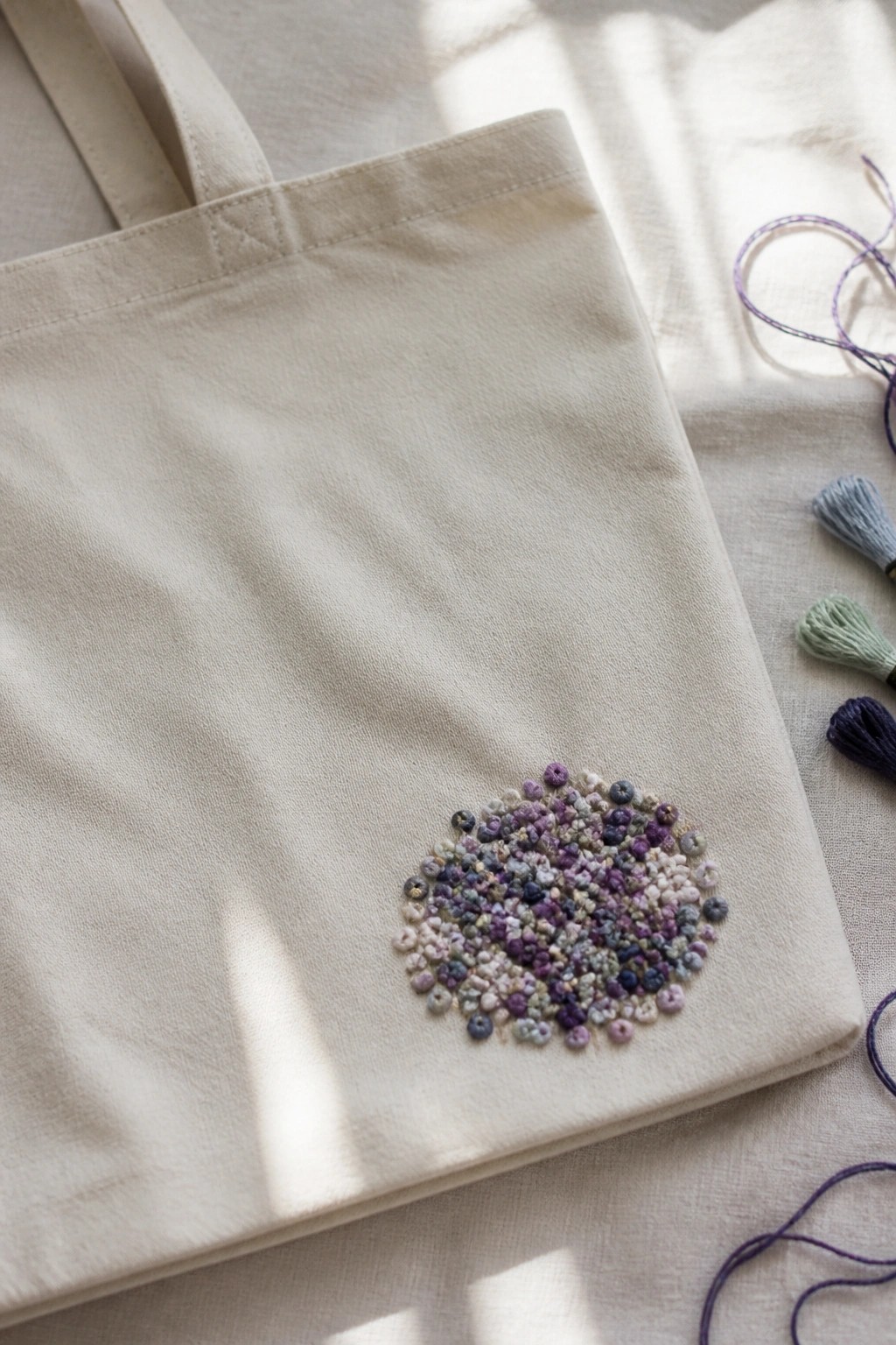 A canvas tote bag with a round cluster of small multicolored embroidery knots on the lower front.