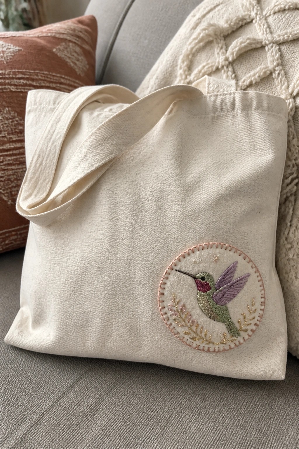 Cream canvas tote bag with embroidered hummingbird in floral circle on couch