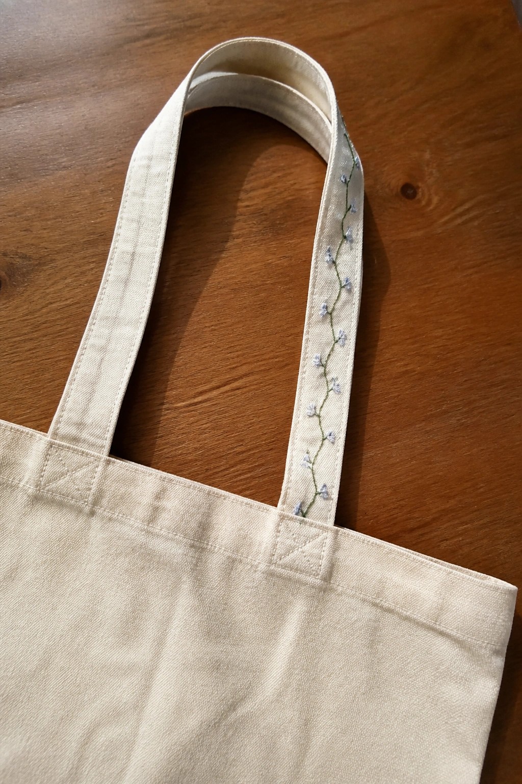 Beige canvas tote bag with blue floral embroidery on handle atop wood table.