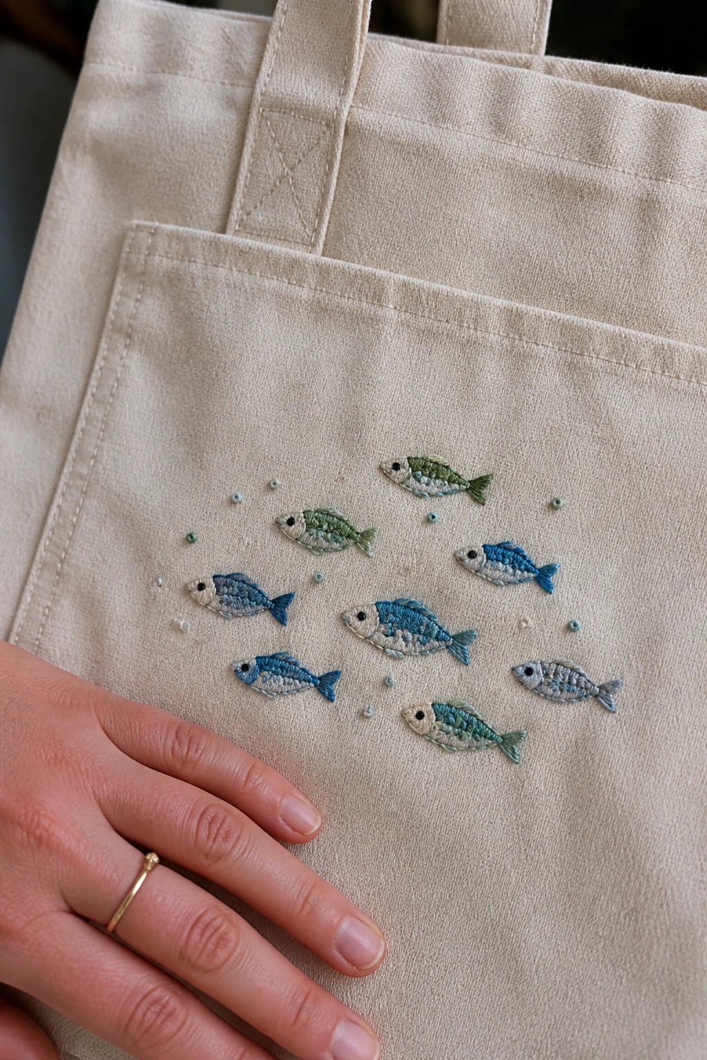 Canvas tote bag with embroidered fish in blue and green threads scattered across the front, small dots around them, and a hand resting on the lower left edge.