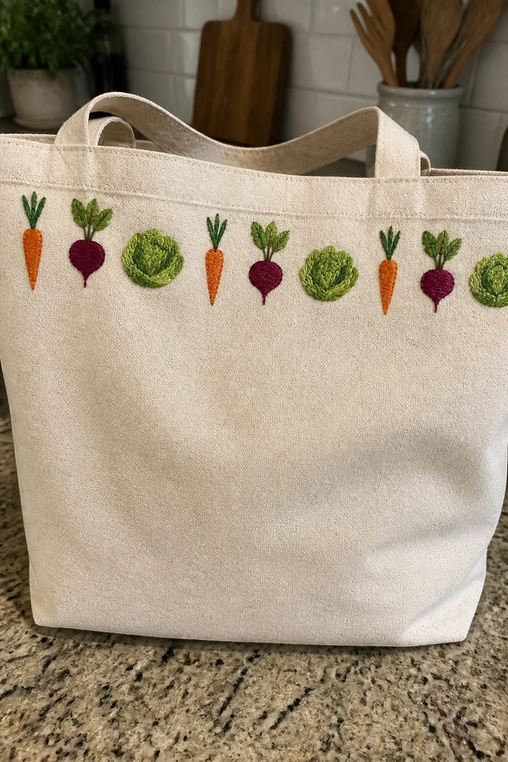 A canvas tote bag with a horizontal row of embroidered carrots, beets, and cabbages along the top front edge.