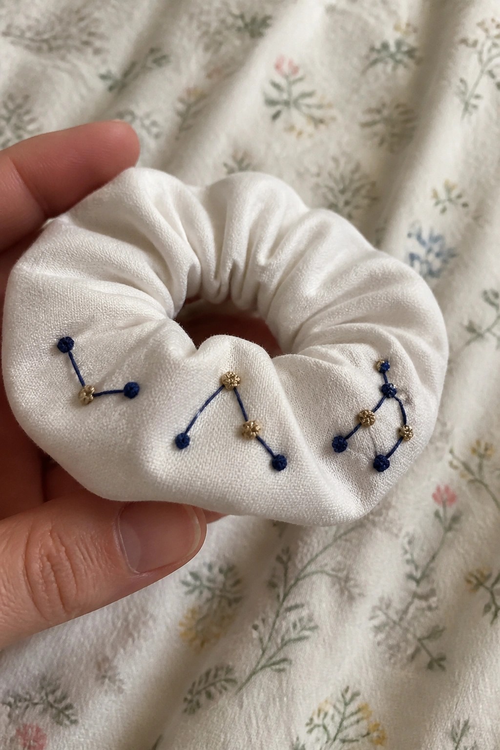 White scrunchie with blue and gold constellation embroidery held on floral fabric.