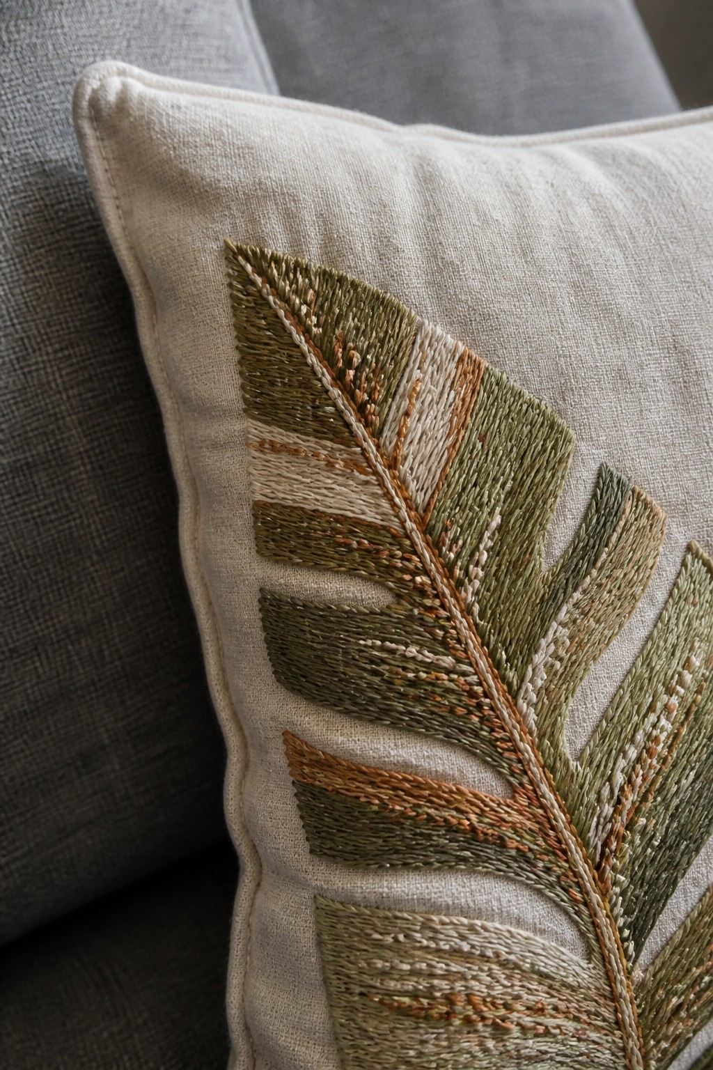 Close-up of a beige cushion with embroidered green and brown leaves in textured stitching.