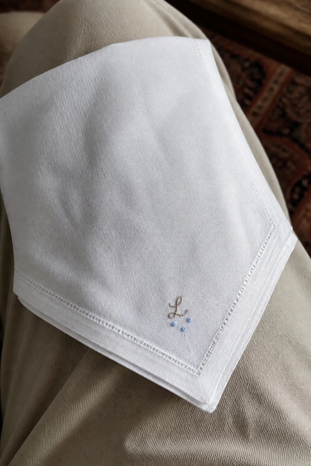 White fabric corner with brown cursive L monogram and three small blue dots embroidered below it.