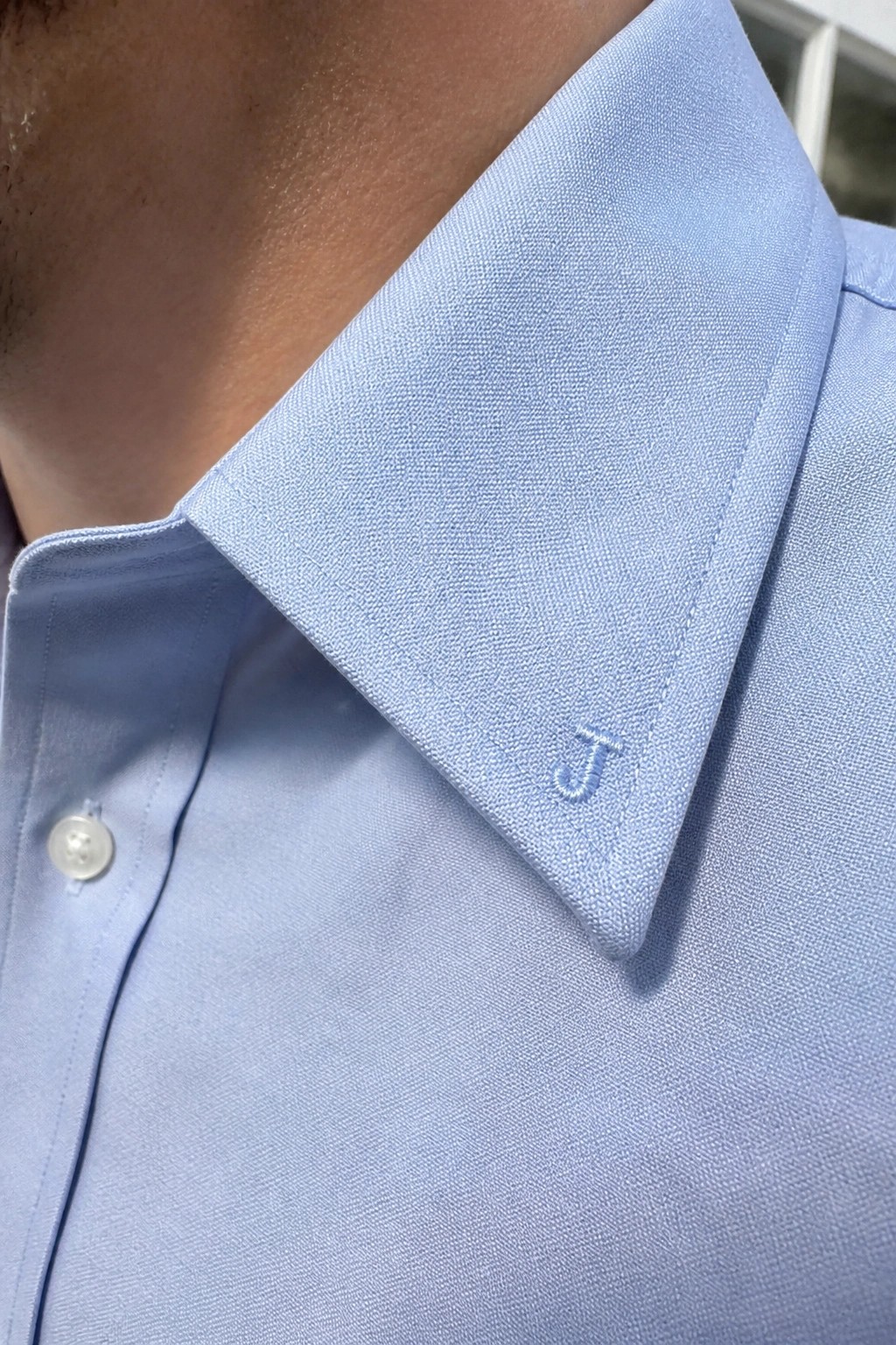 Close-up of a light blue button-up shirt collar with a small embroidered blue letter J on the right tip.