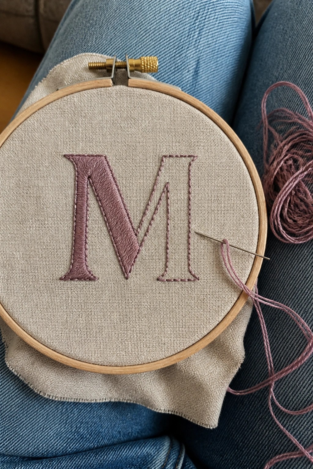 Wooden hoop on denim lap with partially filled purple M on beige fabric.