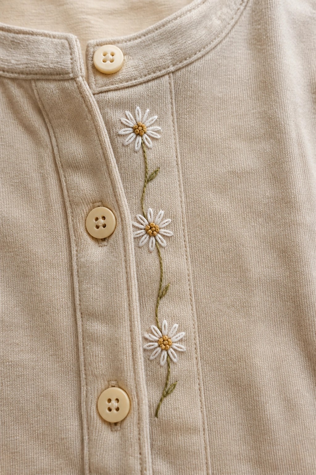 Beige button-up shirt with three white daisies embroidered vertically along the button placket on a green stem.