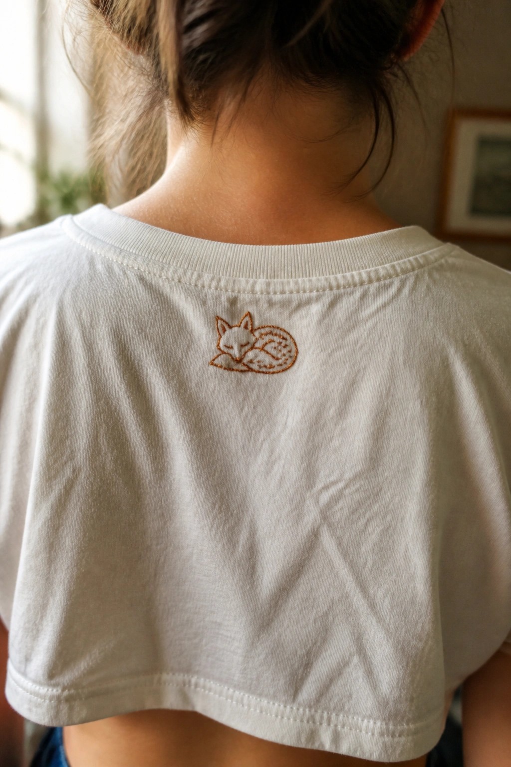 Close-up of the back of a white cropped t-shirt with a small orange embroidered sleeping fox near the neckband.