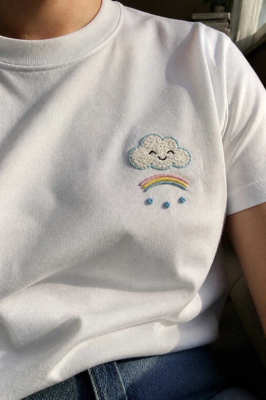 White t-shirt with embroidered smiling cloud, rainbow, and blue raindrops