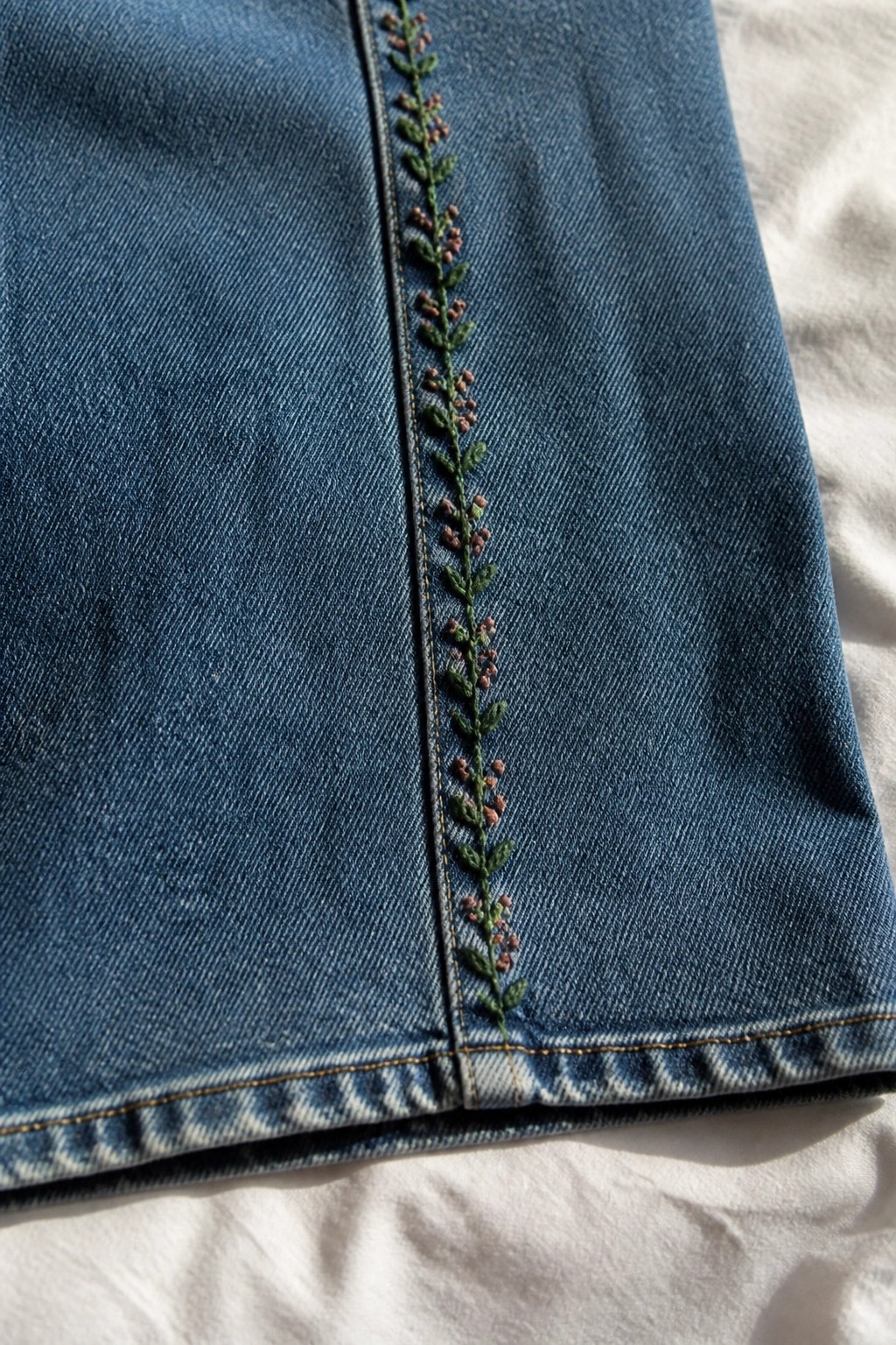 Blue denim with green vine and pink flower embroidery along vertical seam on white surface
