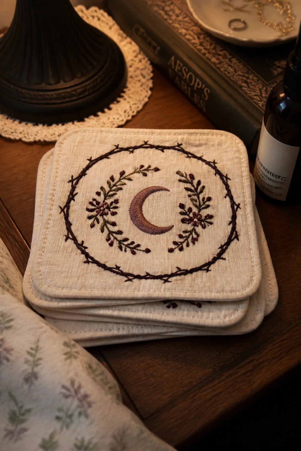 Stack of square fabric coasters embroidered with a crescent moon, floral sprigs, and a thorny circular border.