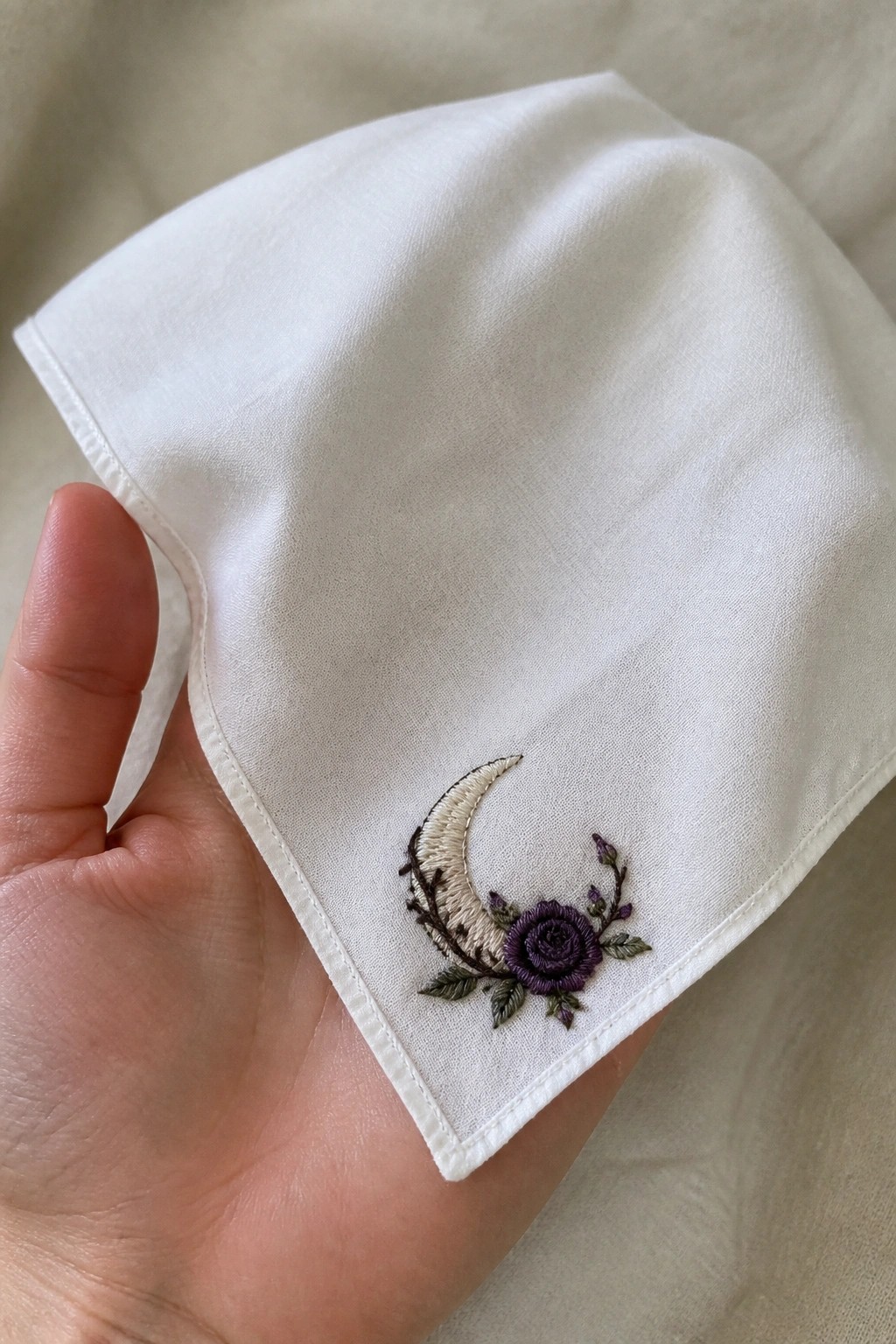 A white fabric corner held in a hand shows an embroidered crescent moon beside a purple rose with leaves and buds.