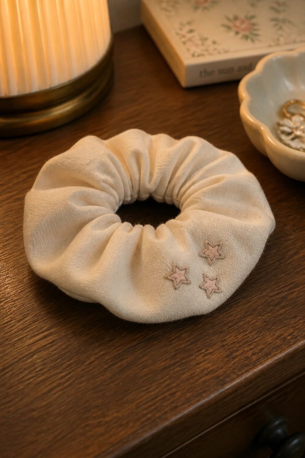 A cream scrunchie with three small embroidered stars in light pink thread resting on a wooden surface.