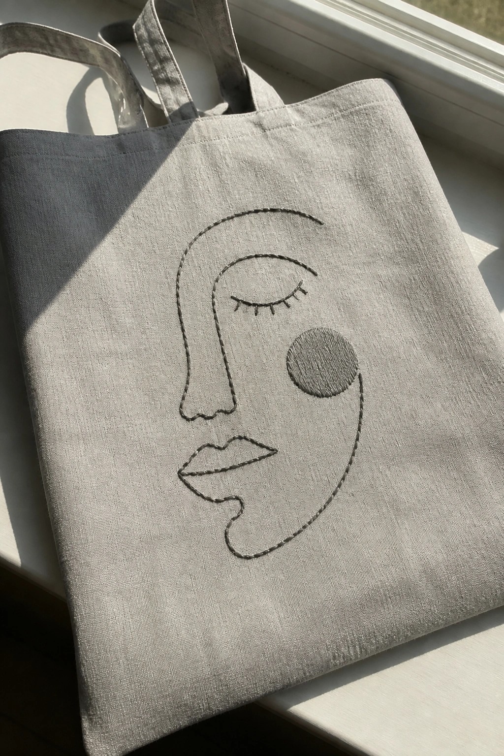 Light gray tote bag with embroidered abstract face on sunny windowsill