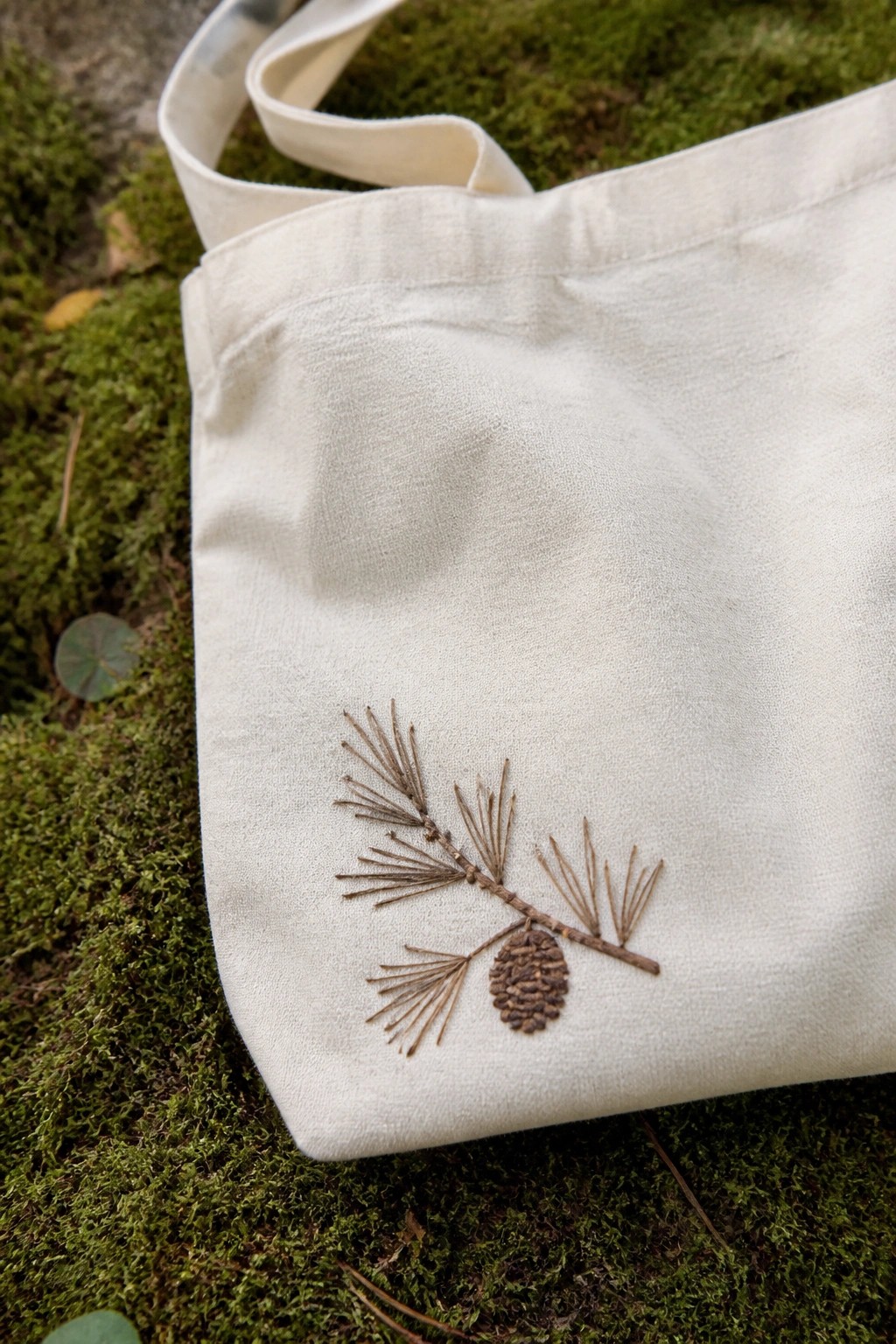 White canvas tote bag with embroidered pine branch and cone on moss.