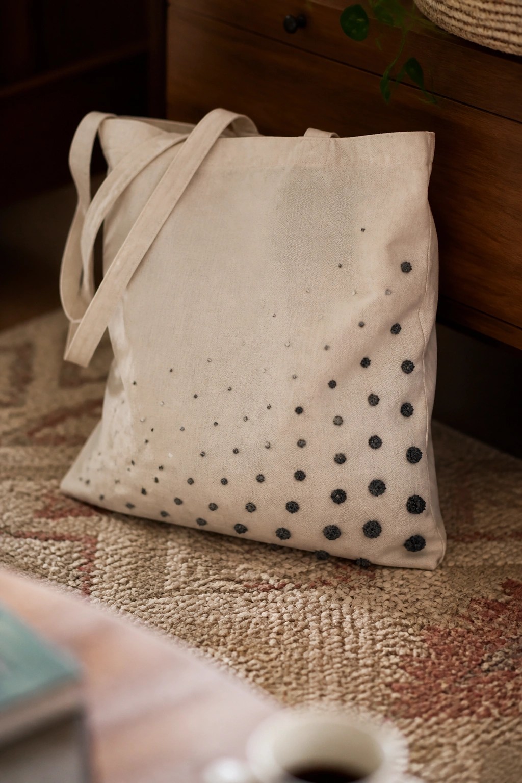 Beige canvas tote bag with scattered black embroidered dots resting on a woven rug.