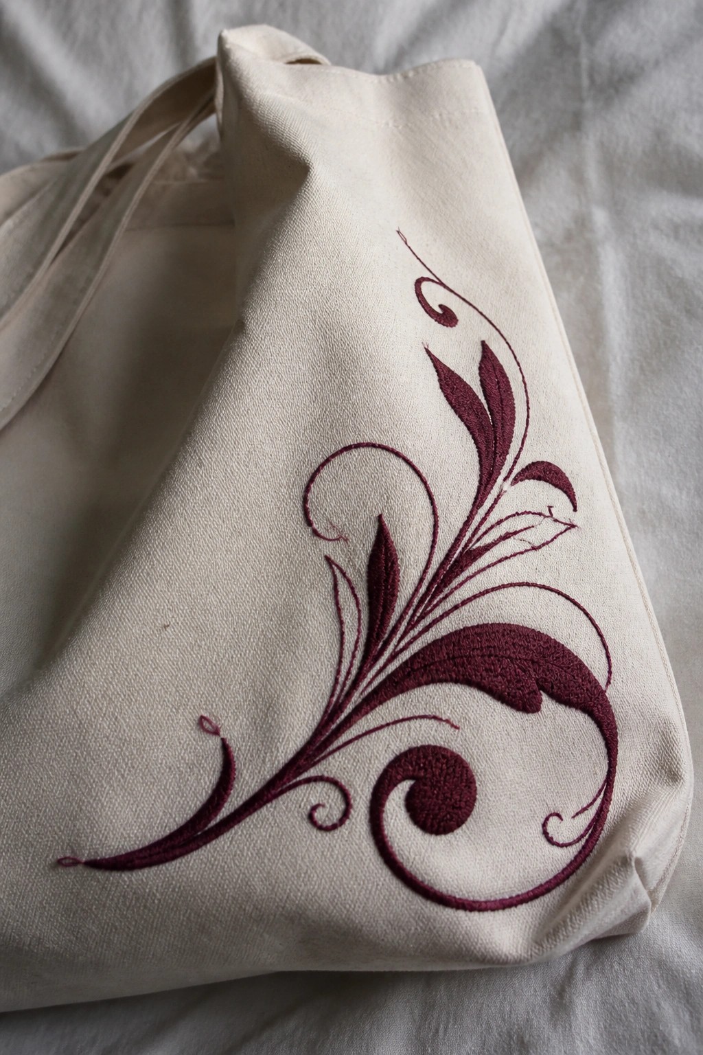 Cream canvas tote bag with maroon embroidered scrolling vine and leaves on the side panel.