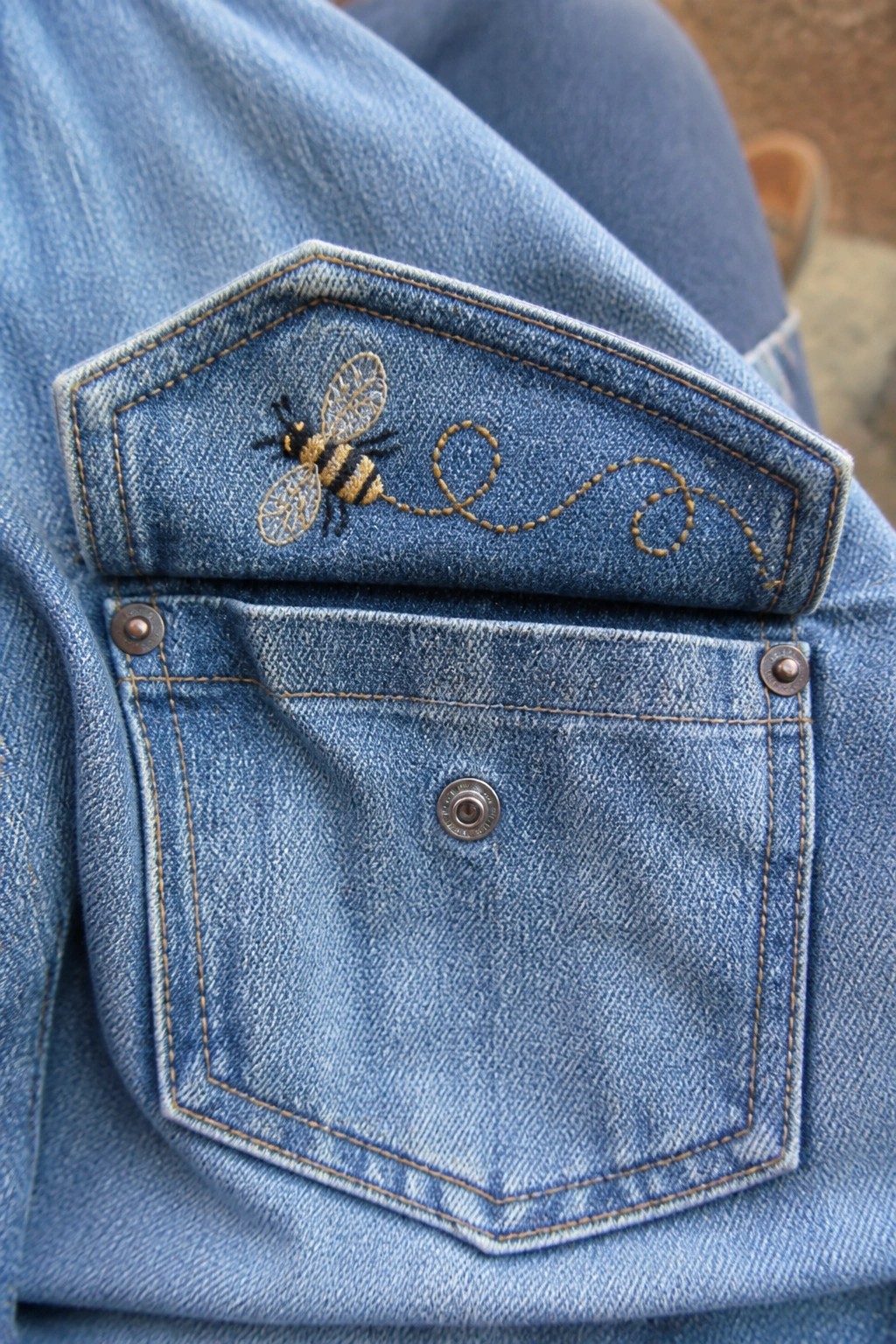 Blue denim pocket with embroidered bee and dotted yellow flight path stitching.