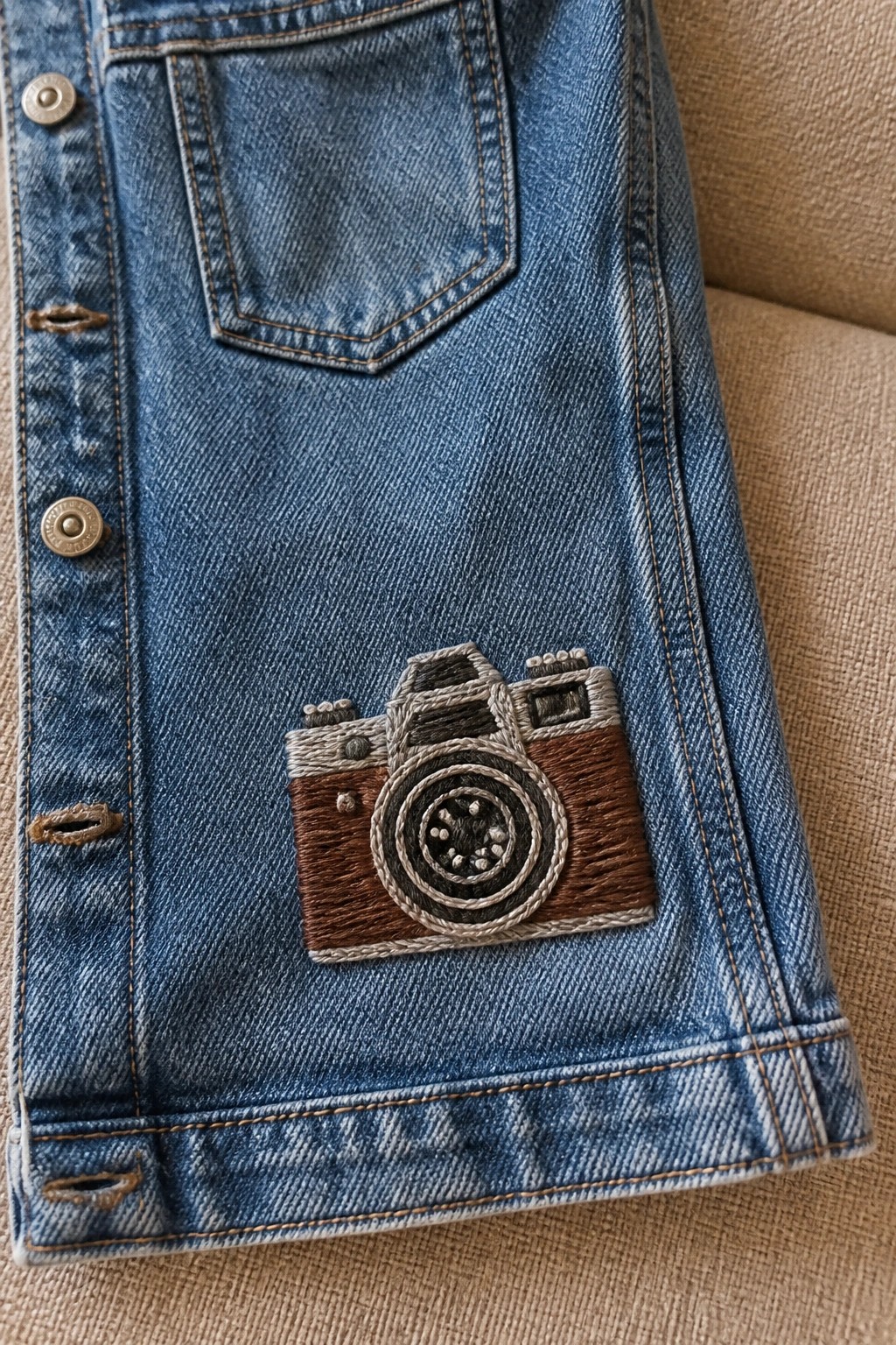 Close-up of blue denim with a brown and gray embroidered camera near the jacket hem and metal buttons.