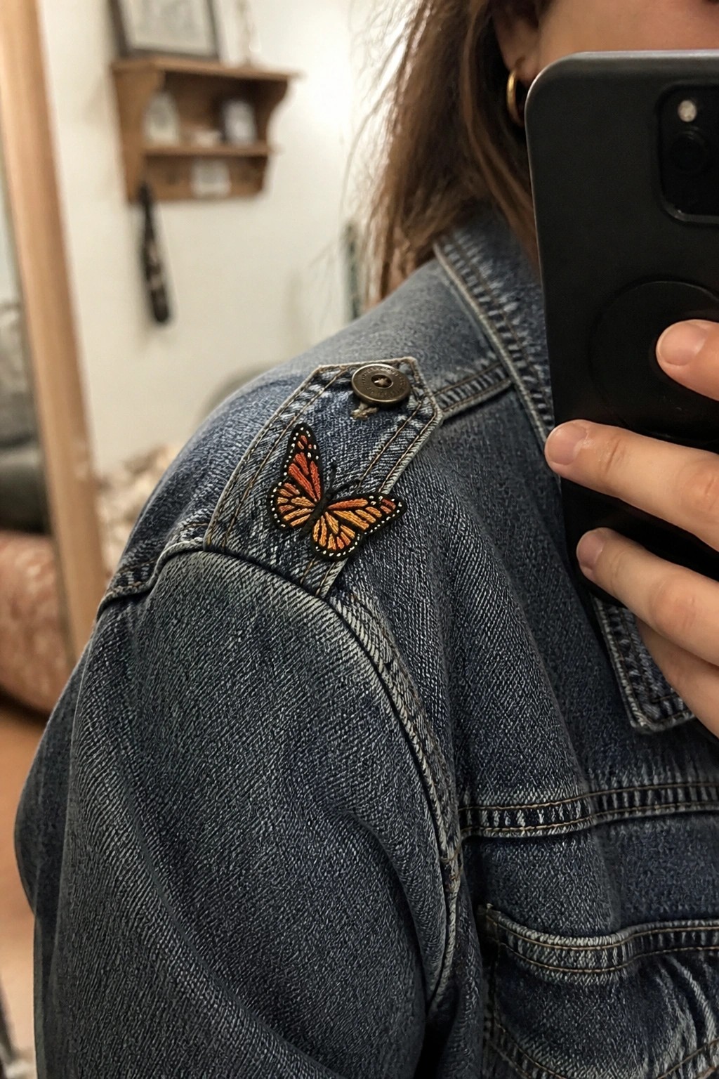 Denim jacket with orange butterfly patch on shoulder, hand holding smartphone