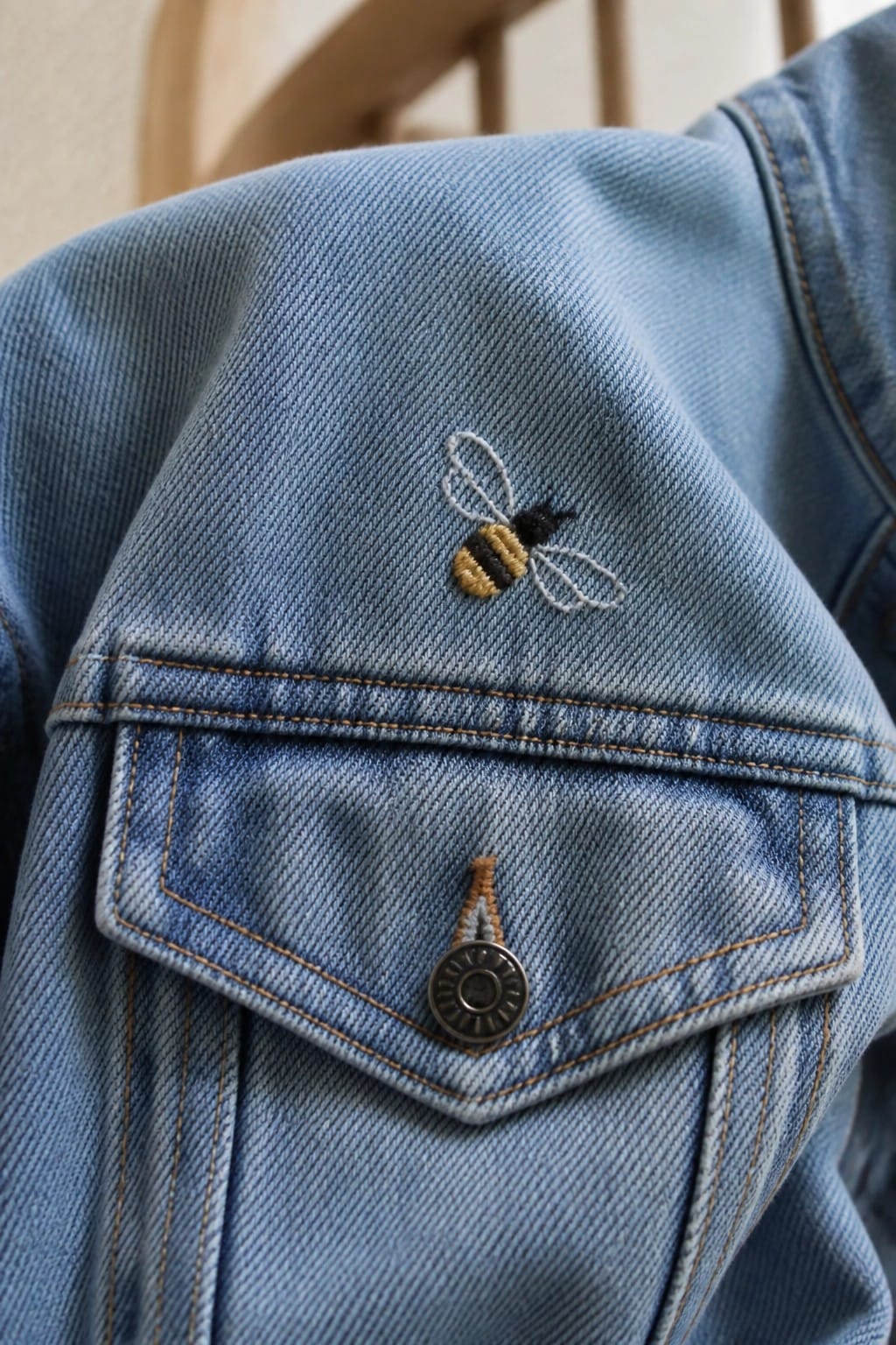 Close-up of blue denim jacket with a small embroidered bee above the pocket flap.