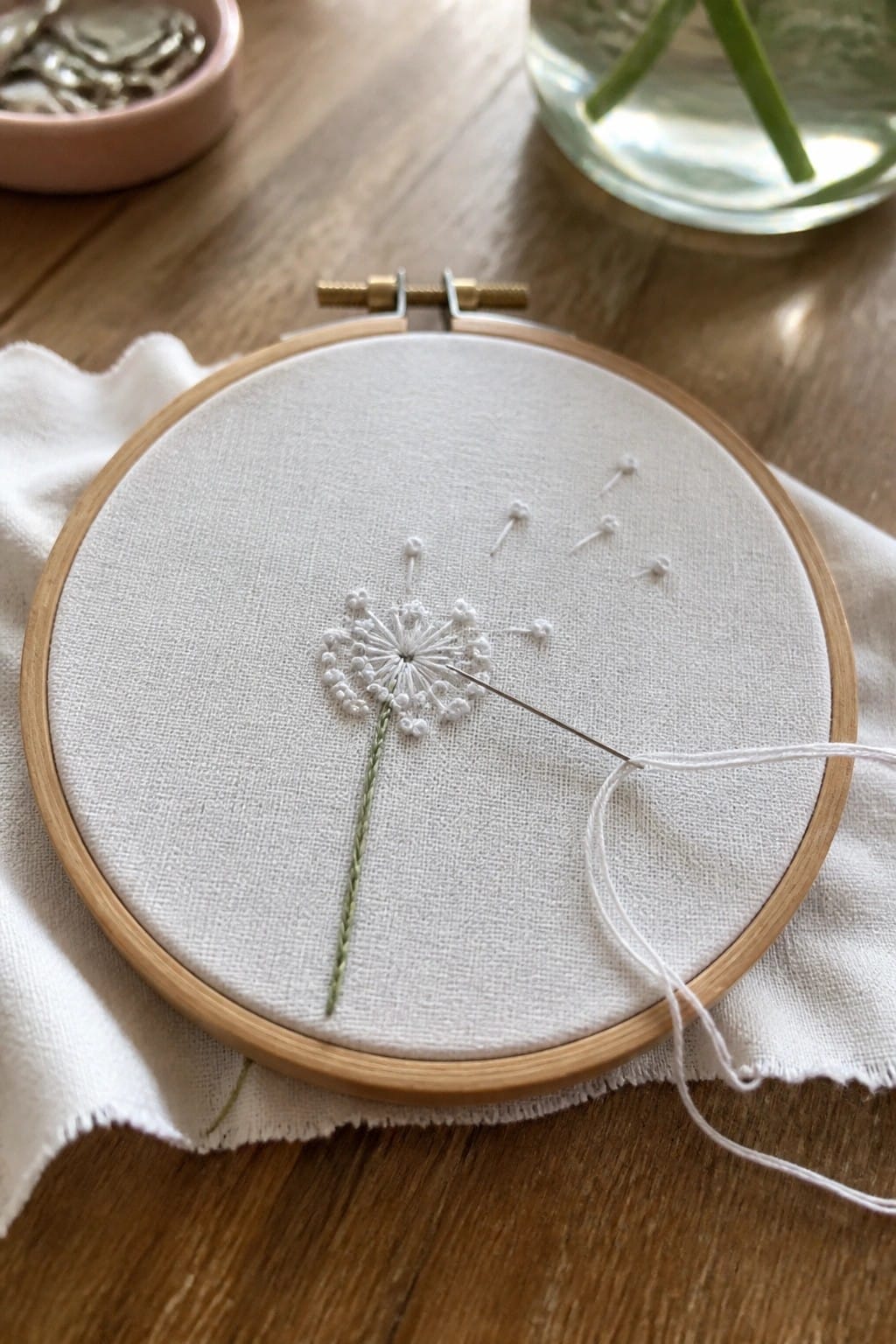 Wooden hoop with white fabric, partial dandelion embroidery, and needle on table.