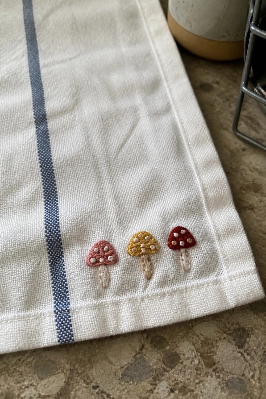 Three small embroidered mushrooms in pink, yellow, and red sit in a row on white fabric next to a blue stripe.