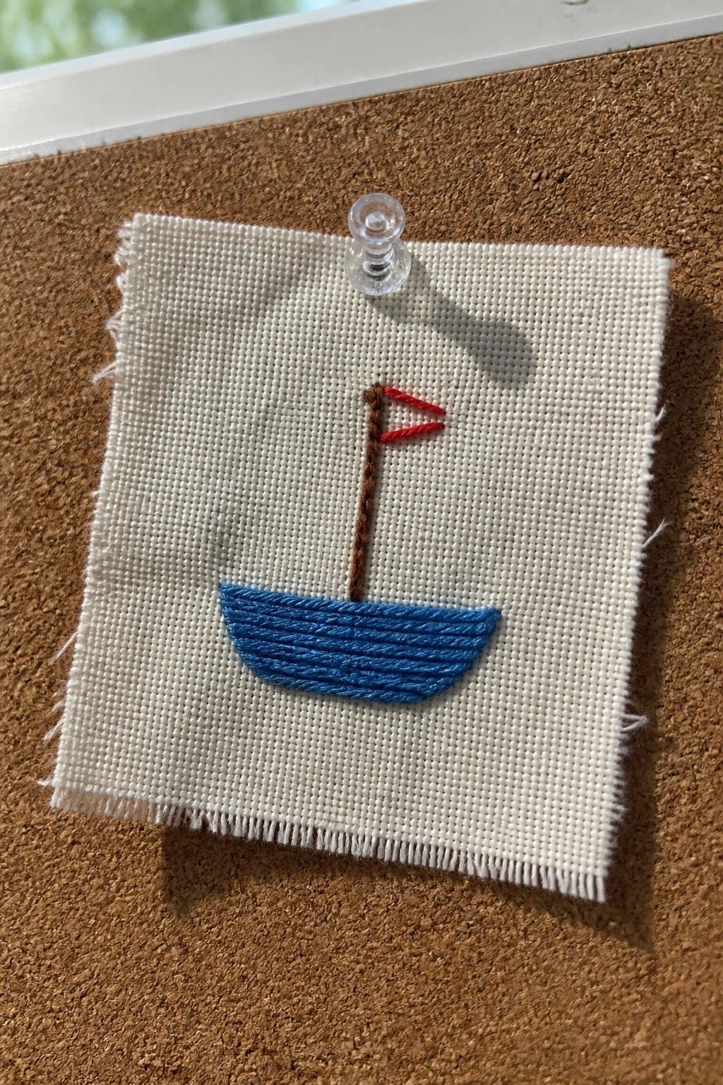 Small square of white fabric with a blue embroidered boat, brown mast, and red flag pinned to corkboard.