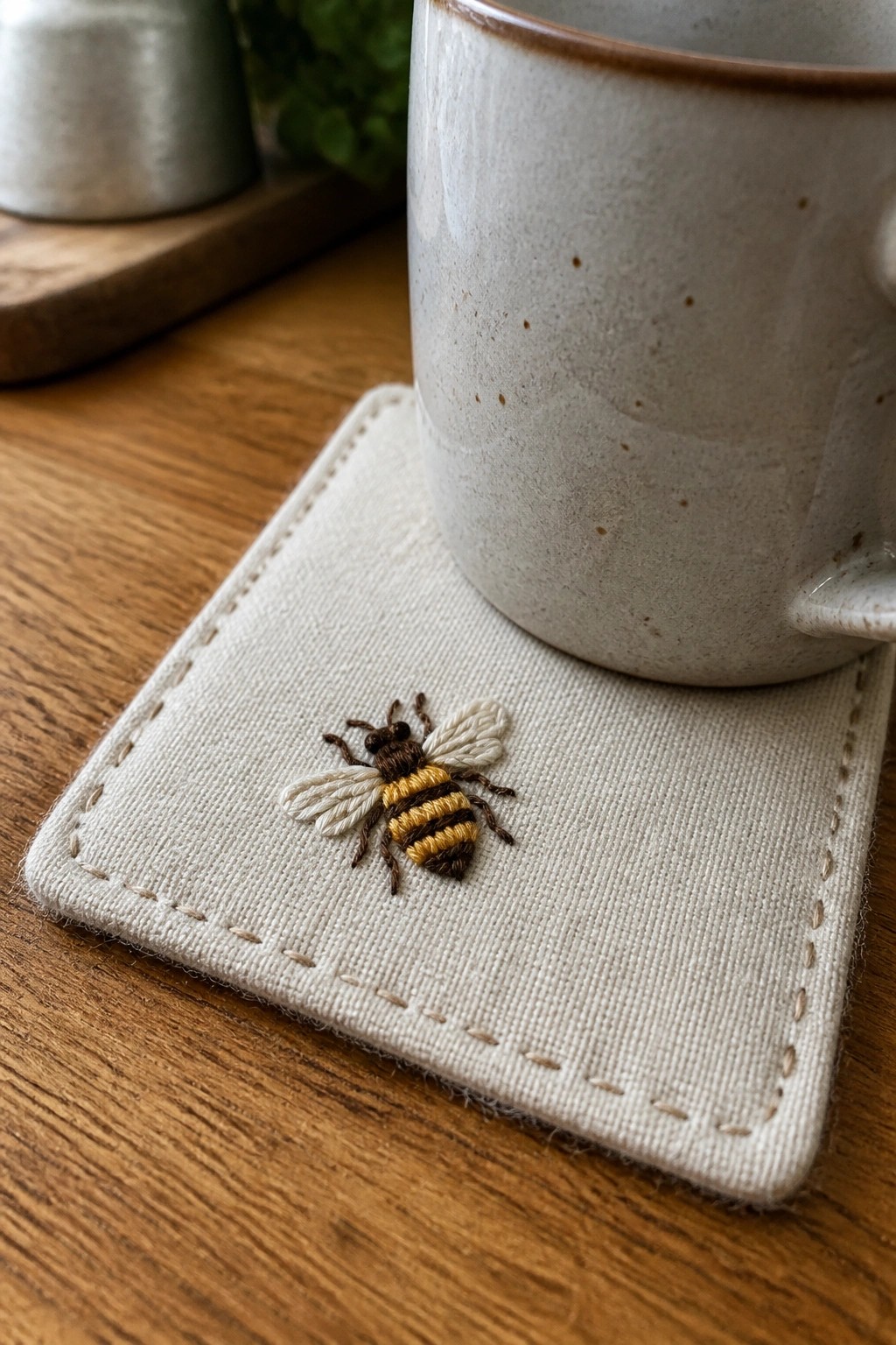Embroidered bee on beige fabric coaster under speckled ceramic mug on wood.