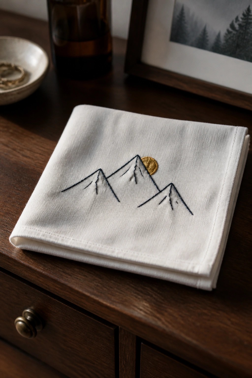 White folded fabric napkin with dark embroidered mountain peaks and a small gold sun centered on the top surface.
