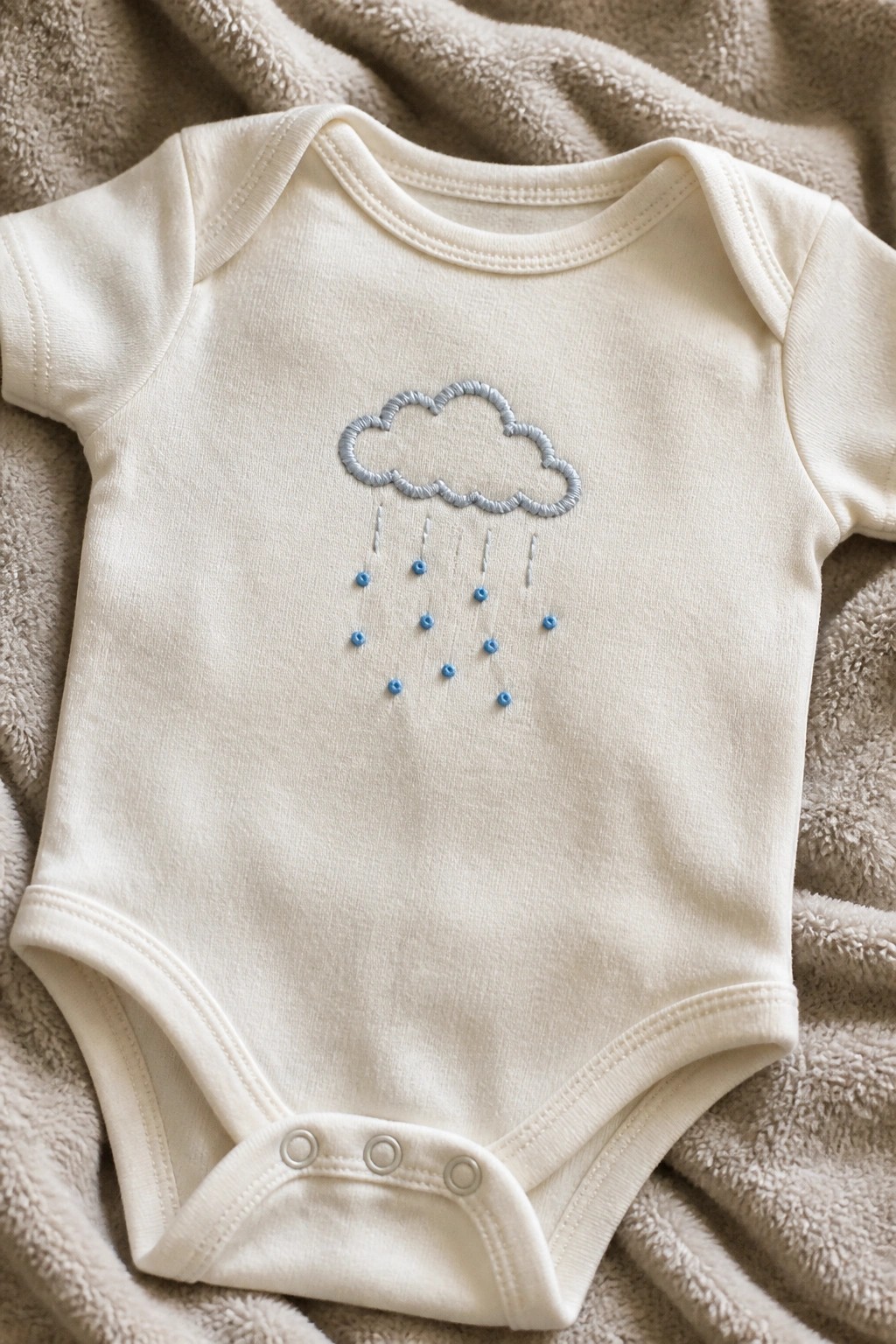 A white baby onesie with a light gray embroidered cloud and blue raindrop dots on the front, laid on a beige blanket.