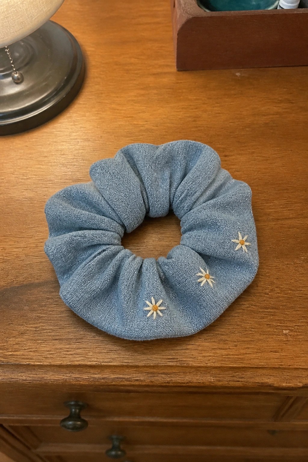 A light blue scrunchie embroidered with three small daisy flowers resting on a wooden table.