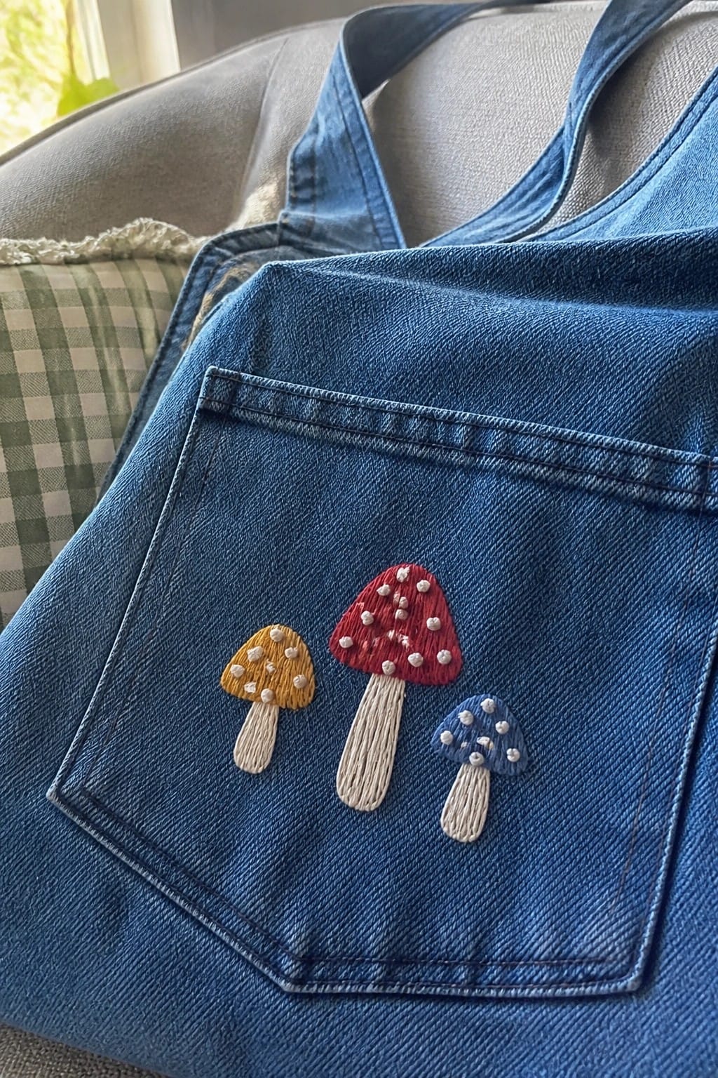 Denim tote bag pocket with three small embroidered mushrooms in yellow, red, and blue.
