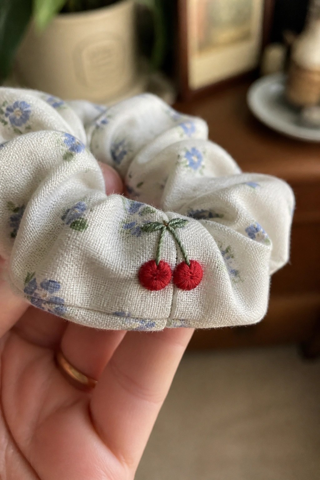 Hand holding white floral scrunchie with red cherry embroidery on fabric.