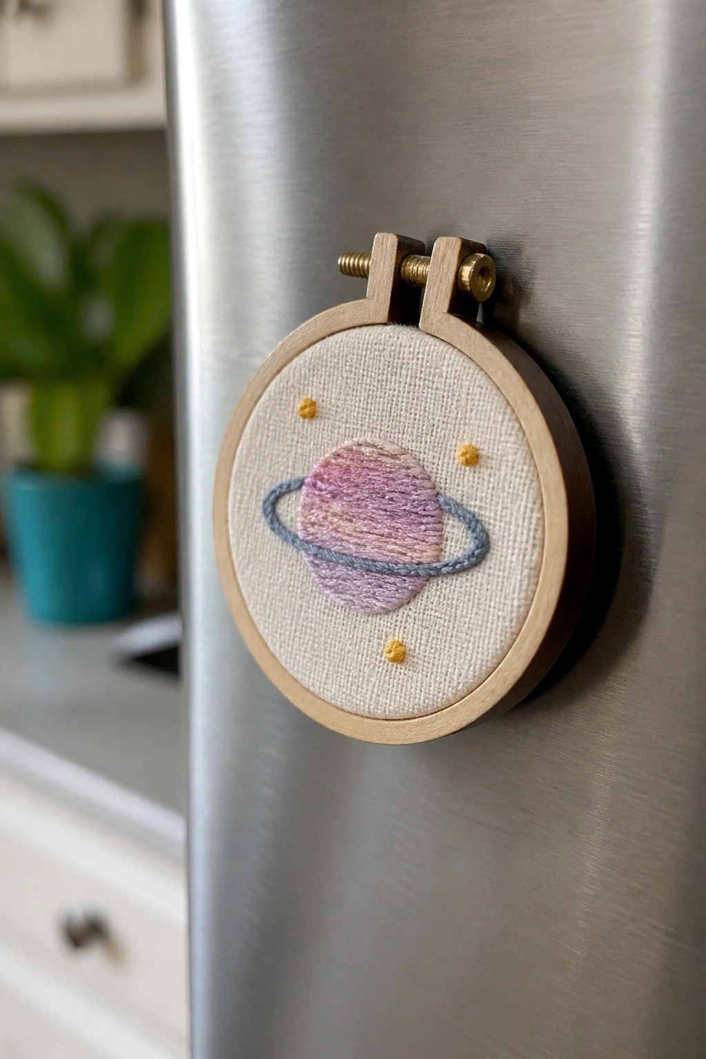 Miniature embroidery hoop with pink-purple planet and blue rings on stainless fridge.