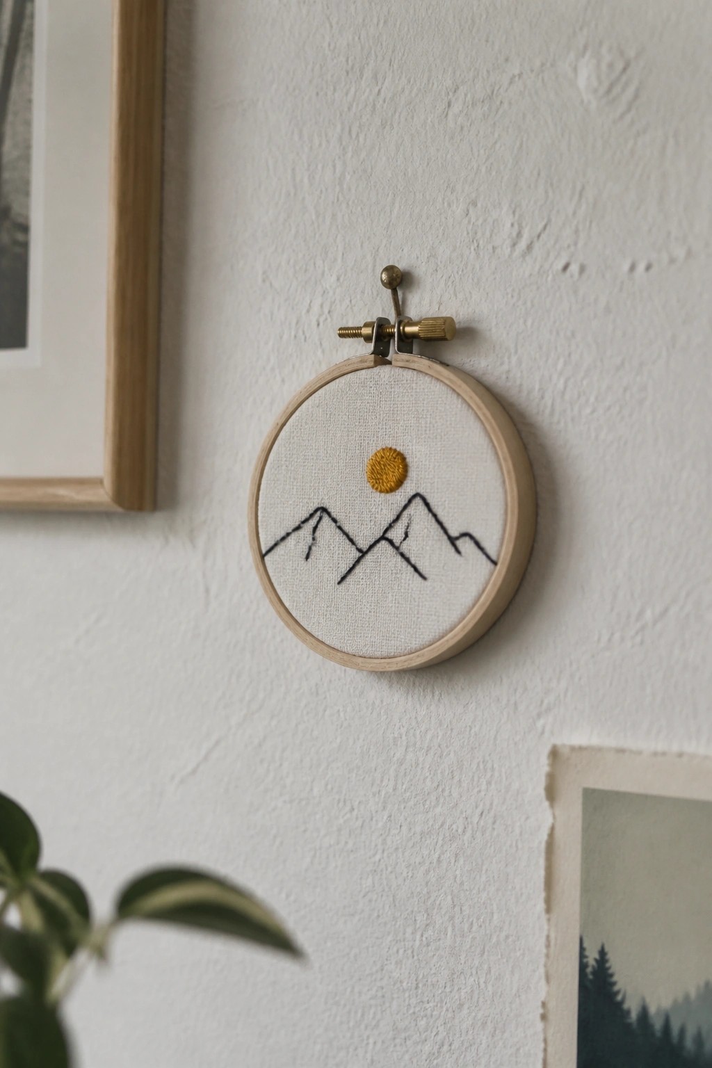 A small embroidery hoop with black mountain outlines and a yellow sun on white fabric hanging on a wall.