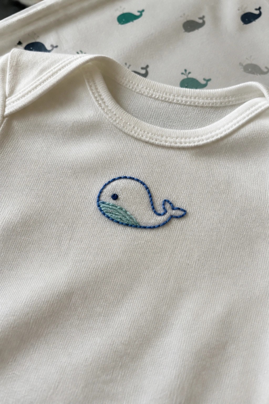 Small blue embroidered whale on the chest of a white baby onesie.