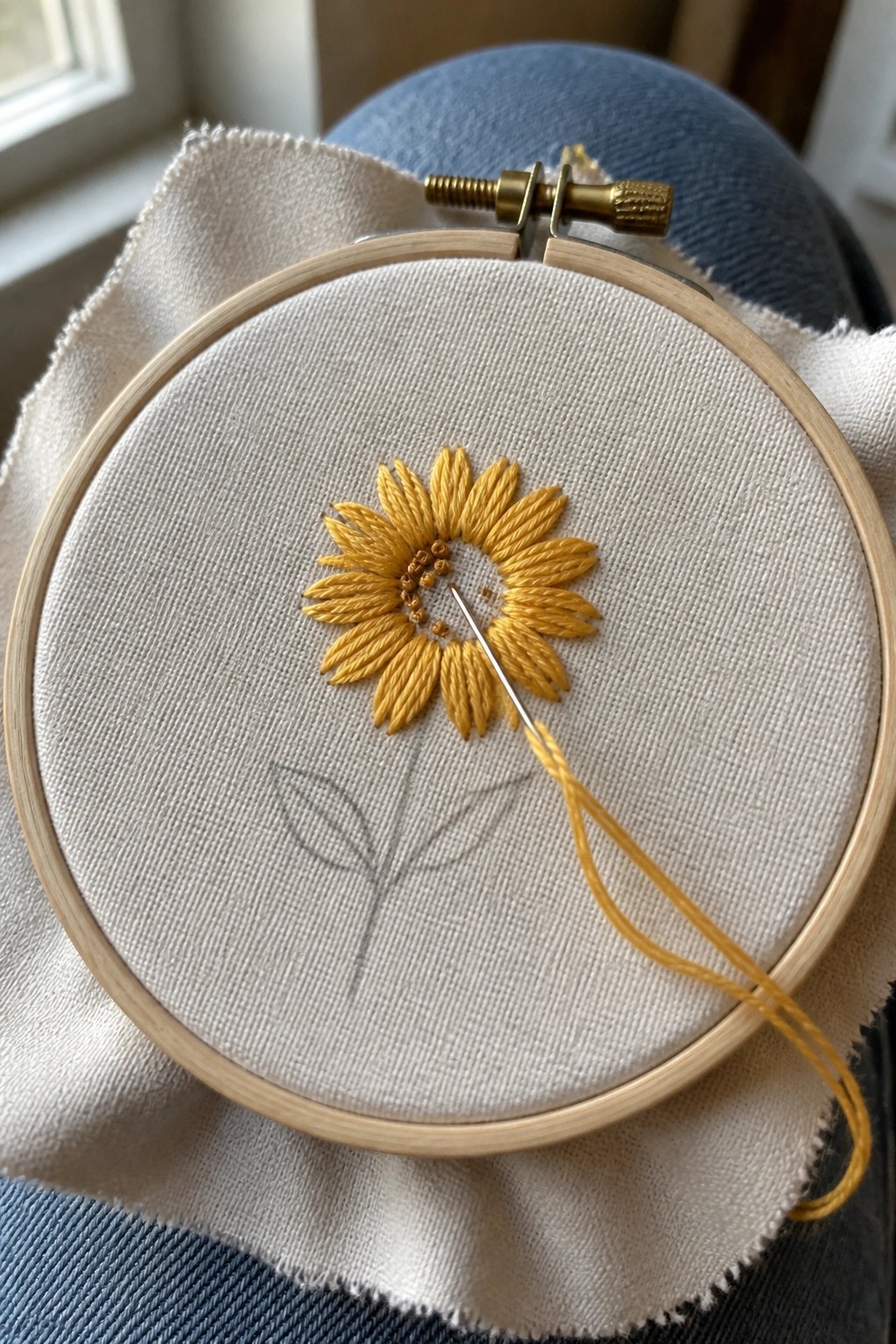 A small wooden embroidery hoop holds white fabric with a yellow sunflower stitched in the center and a stem with leaves sketched below, needle and thread resting on the fabric.