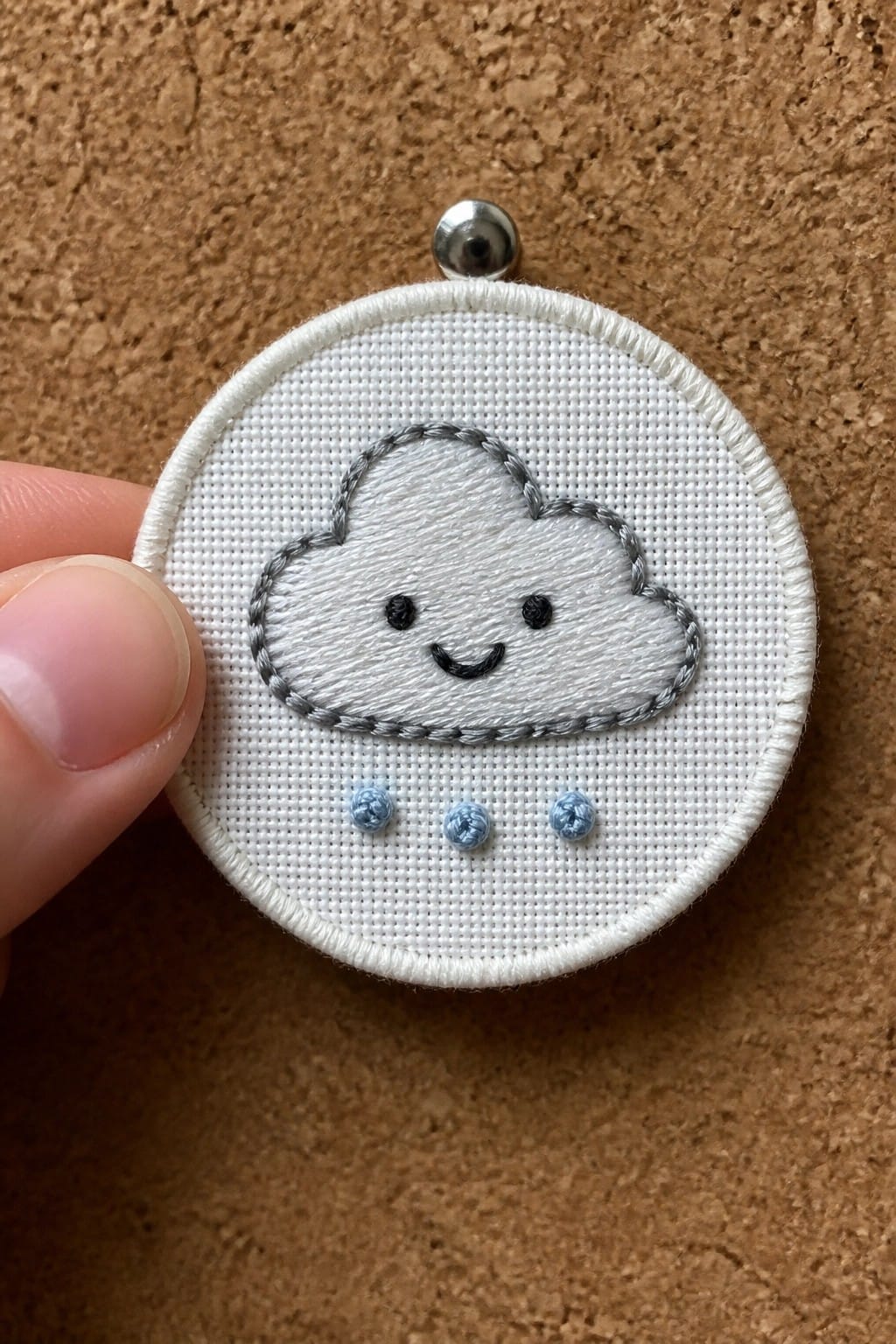 Small round embroidery hoop with a white cloud outlined in gray thread, black eyes and smile, and three blue knotted raindrops below on white fabric.