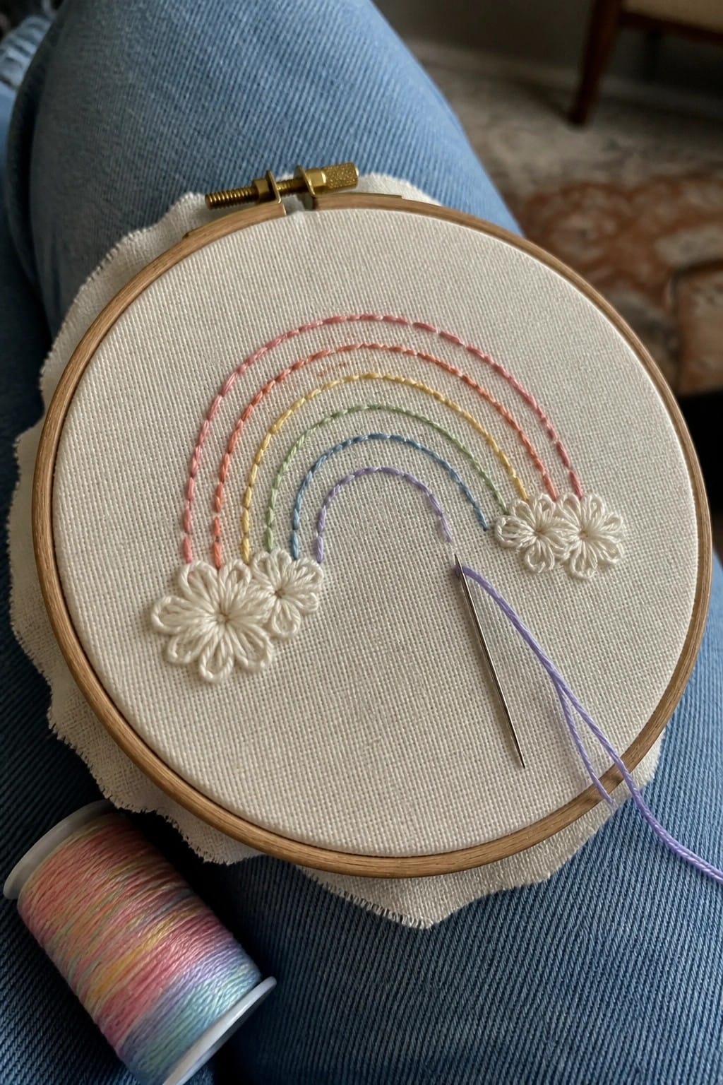 Colorful rainbow embroidery hoop on blue jeans with needle and thread spool nearby.