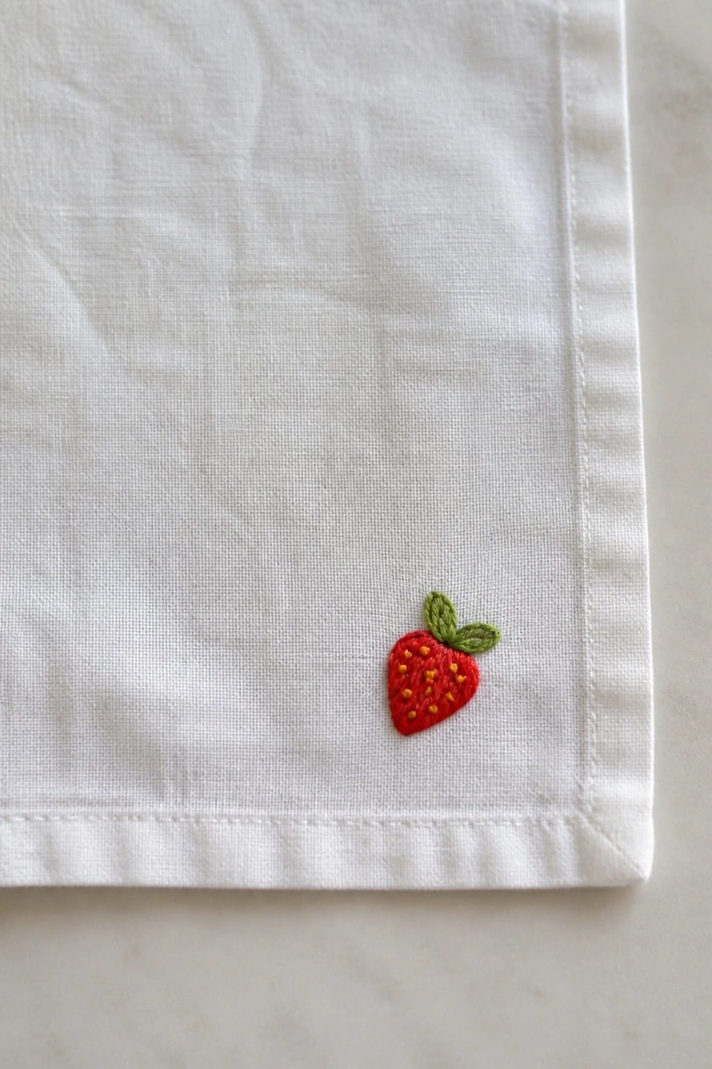 A small red strawberry with green leaves and yellow seeds is embroidered on the corner of a white fabric napkin.