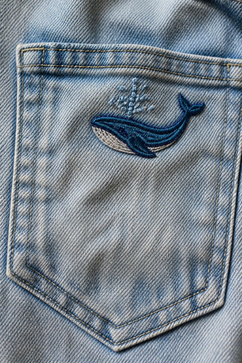 A blue embroidered whale on a light denim jeans pocket with small water spray stitches above it.