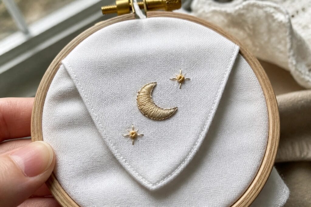 Hand holding an embroidery hoop with white fabric showing a stitched crescent moon and two stars on a triangular section.